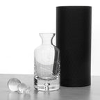 Clear glass carafe with handle and stopper next to a black cylindrical container on a white background