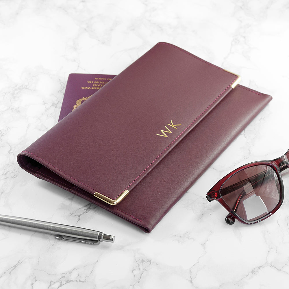 Maroon passport holder with initials, pen, glasses, and passport on a marble surface