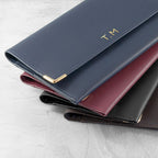 Three leather wallets in different colors stacked on a light gray surface with 'T.M.' branding.