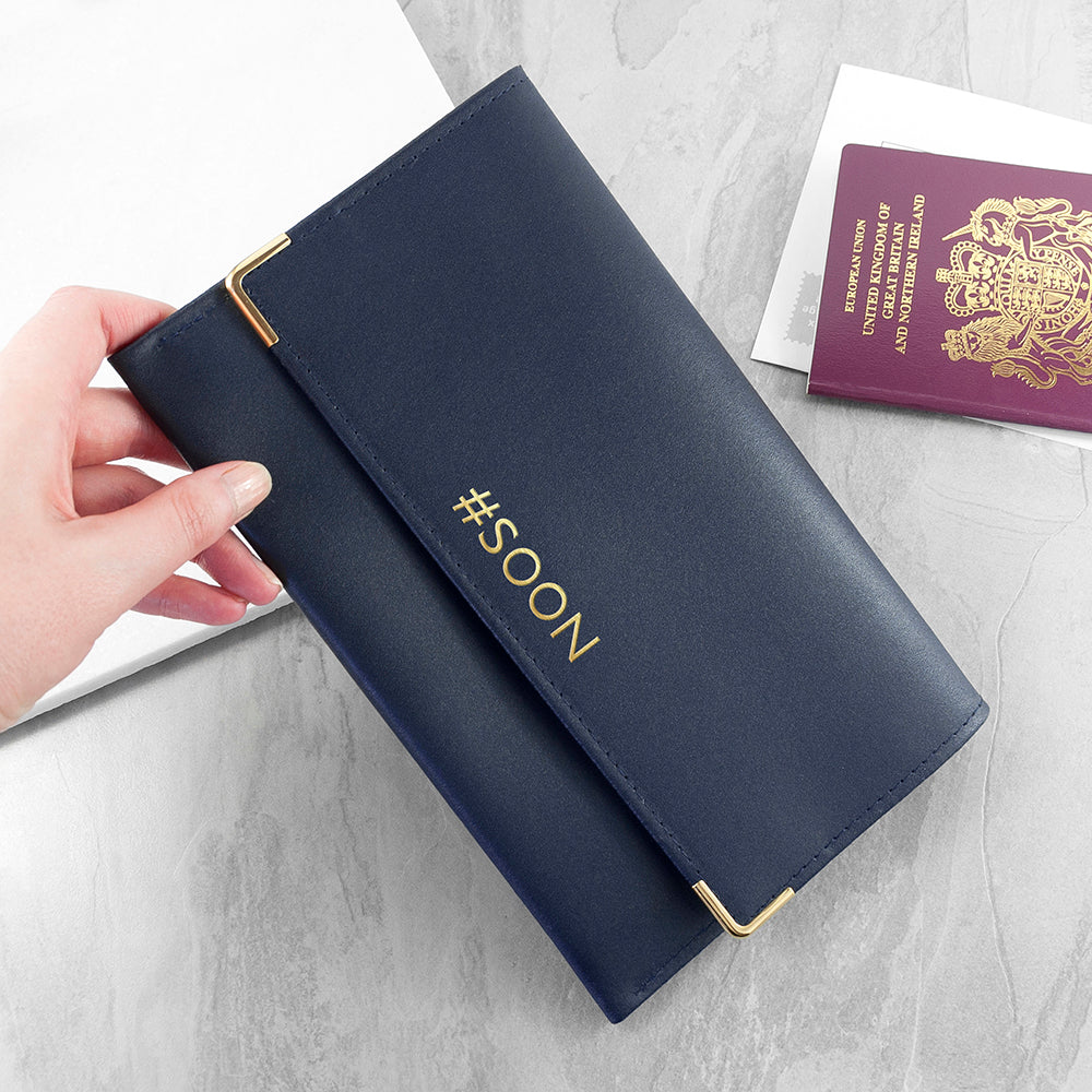 Navy blue passport holder with '#SOON' text held by a hand, on a light gray surface with a passport.