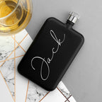 Black flask with 'Jack' engraving on a marble surface with a glass of whiskey.