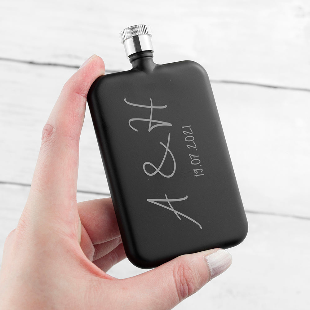 Black hip flask with engraved initials and date held by a hand on a light background