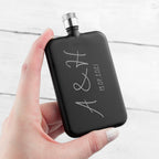 Black hip flask with engraved initials and date held by a hand on a light background
