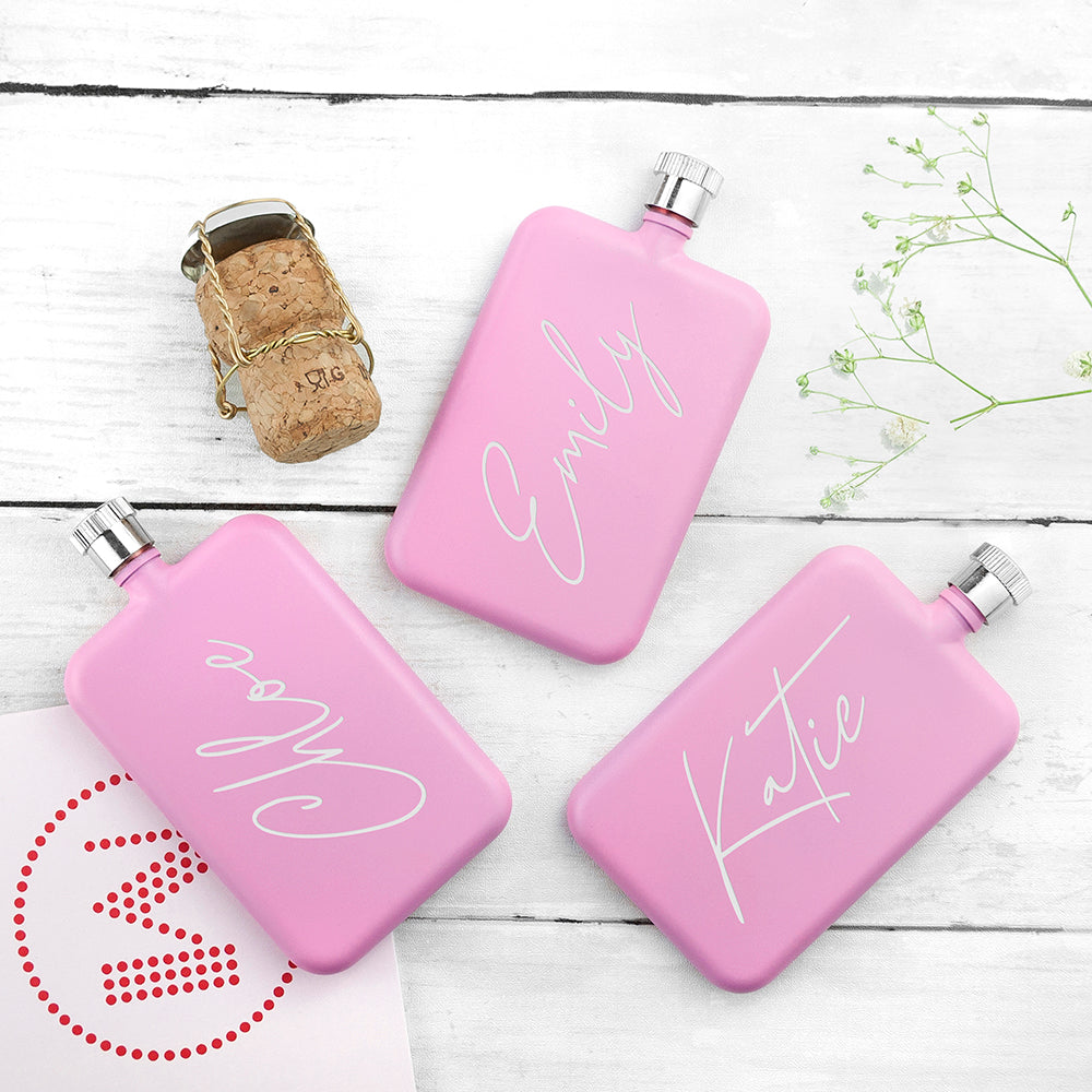 Three pink flasks with names 'Emily', 'Lola', and 'Katie' on a wooden surface.