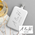 White flask with engraved initials and date on a marble surface with a glass of white wine.