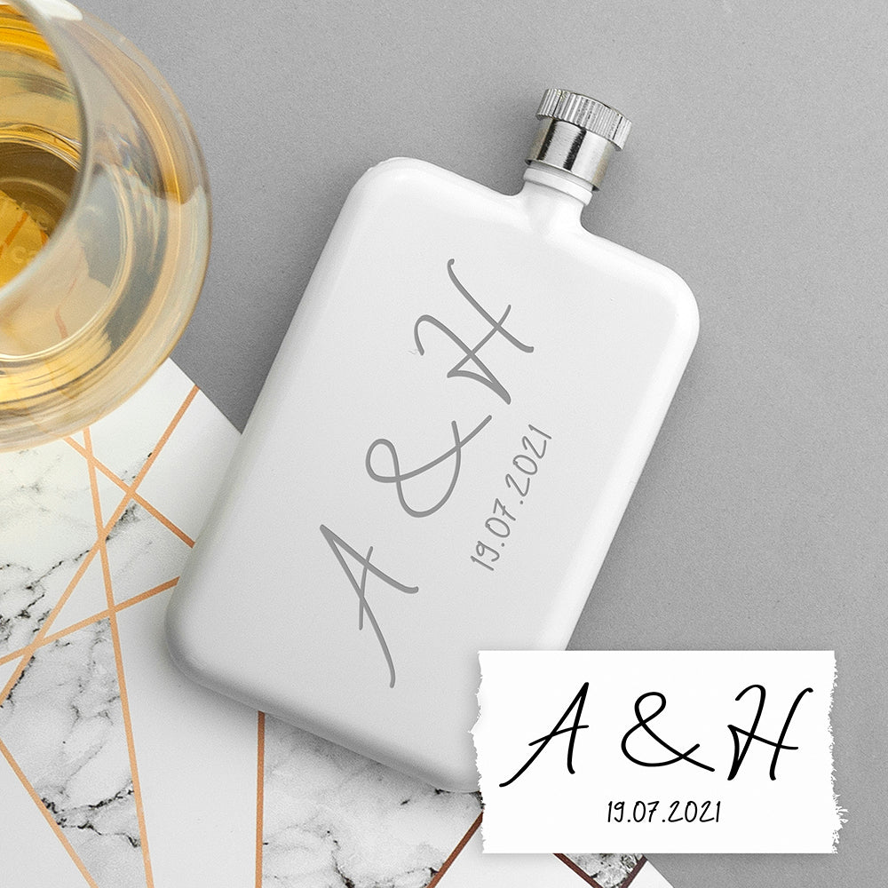 White flask with engraved initials and date on a marble surface with a glass of white wine.