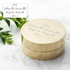 Round brass box with engraved message on a marble surface with green leaves.