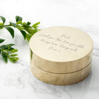 Round brass box with engraved text on a marble surface with green leaves.