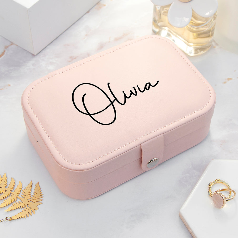Pink jewelry box with 'Olivia' engraving on a marble surface with gold jewelry.