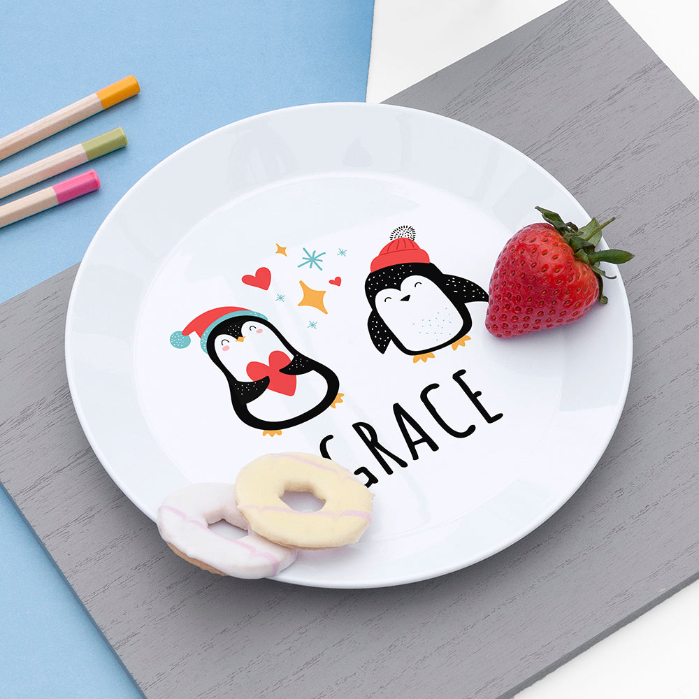 White plate with cartoon penguins and the name 'Grace', placed on a gray surface with a strawberry and donut.