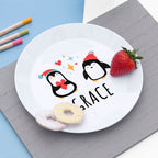 White plate with cartoon penguins and the name 'Grace', placed on a gray surface with a strawberry and donut.