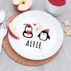 Round white plate with cartoon penguins and 'ALFIE' text on a cork placemat, surrounded by apples and a cup.