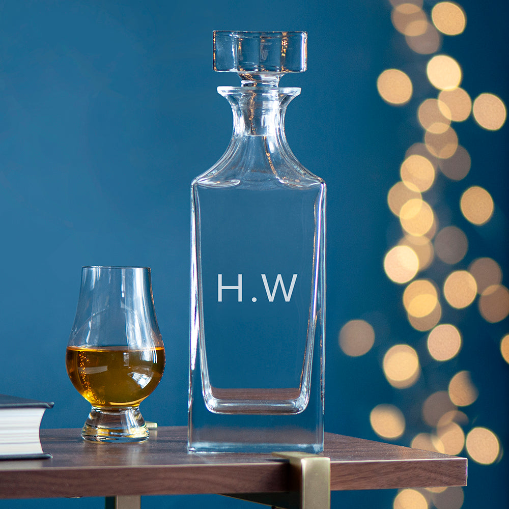 Clear glass decanter with 'H.W.' initials next to a filled glass on a wooden surface with a blurred light background.