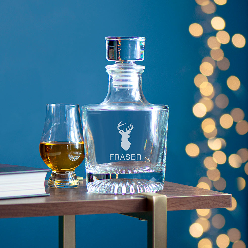 Clear glass decanter with 'FRASER' engraving on a wooden table against a blue background with bokeh lights.