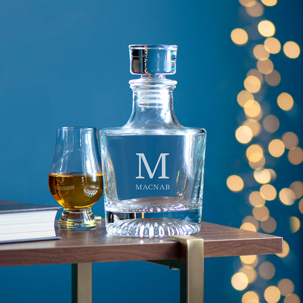 Clear glass decanter with 'M' monogram and glass on a wooden table against a blue background with bokeh lights.