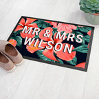 Floral doormat with 'MR & MRS WILSON' text on a white floor with shoes and a plant.