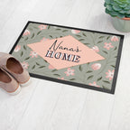 Decorative doormat with floral pattern and 'Nana's Home' text on a light wooden floor.