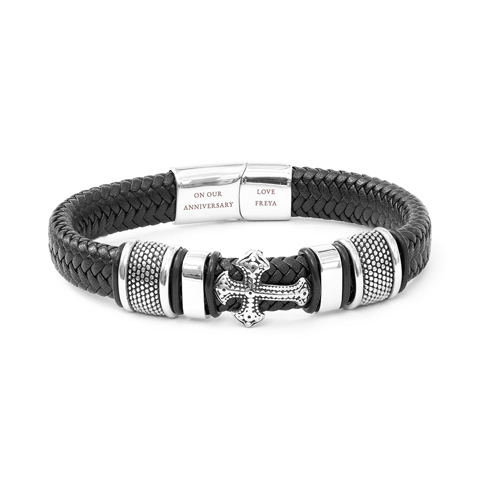 Black braided leather bracelet with silver cross charm and engraved tag on a white background