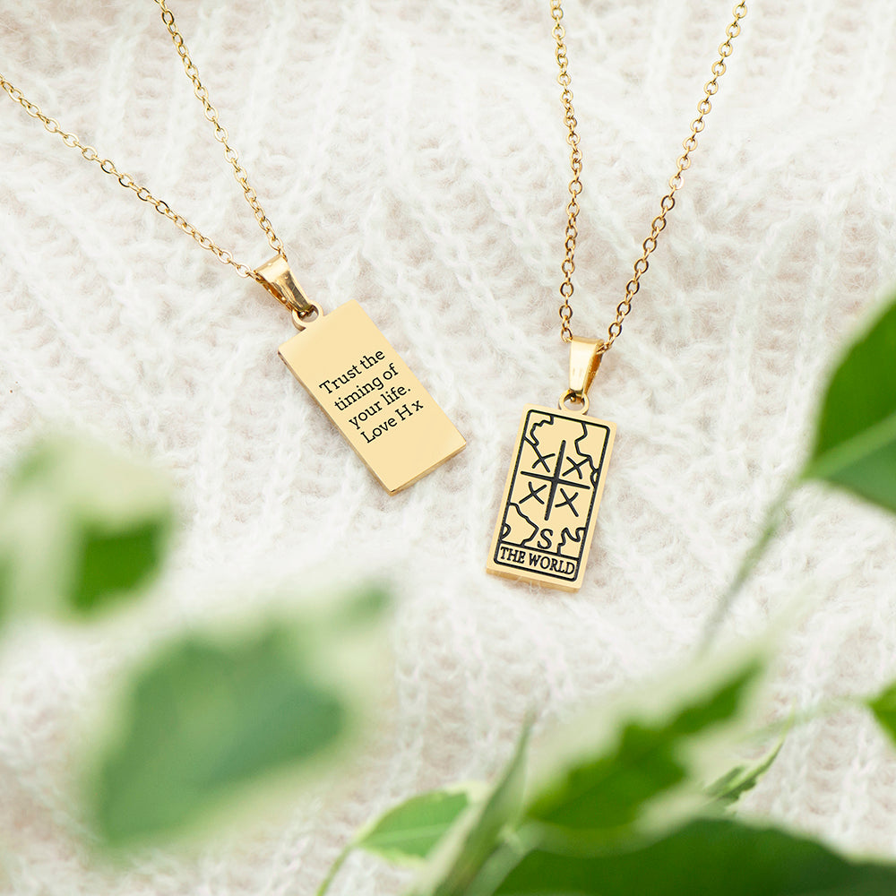 Two gold necklaces with pendants on a textured surface with green leaves in the background