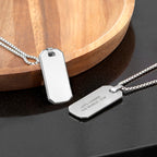 Two silver dog tag necklaces on a dark surface with a wooden circular object in the background.