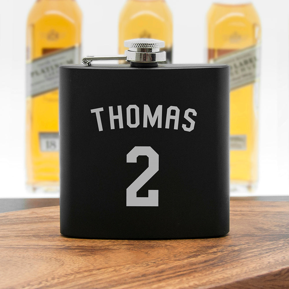 Black flask with 'THOMAS 2' printed on it, placed on a wooden surface with blurred bottles in the background.