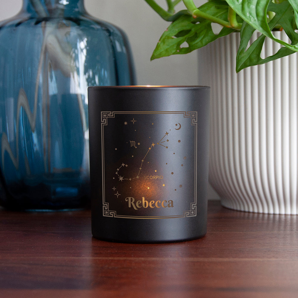 Black candle with 'Rebecca' and zodiac design on a wooden surface with decorative items.