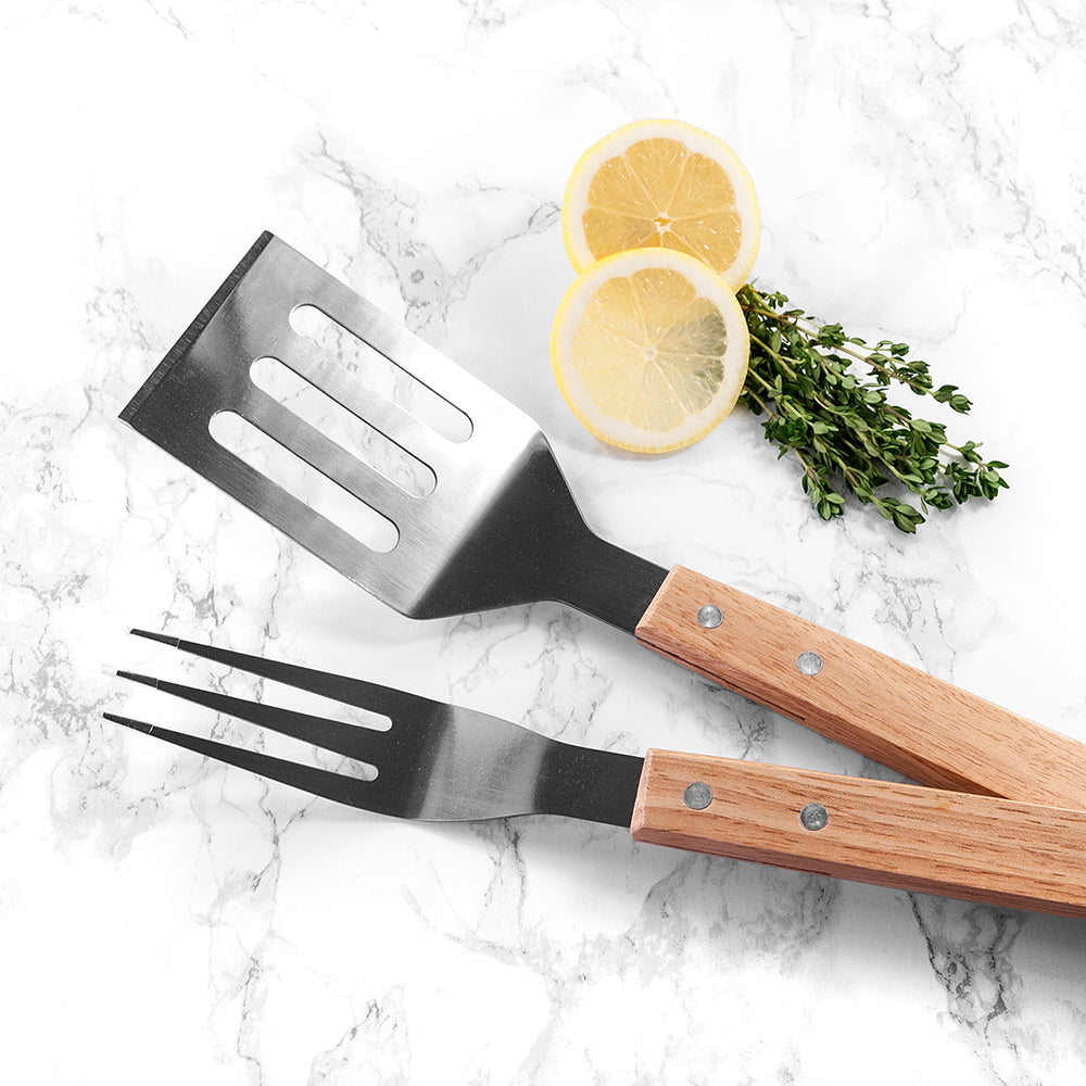 Barbecue utensils with wooden handles on a marble surface with lemon slices and herbs.
