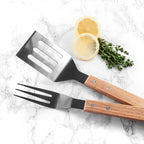 Barbecue utensils with wooden handles on a marble surface with lemon slices and herbs.