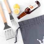 Barbecue utensils, lemons, and a beer bottle on a white surface with a gray apron.