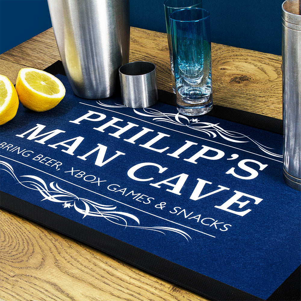 Personalized bar mat with 'PHILIP'S MAN CAVE' text on a wooden surface with bar accessories.