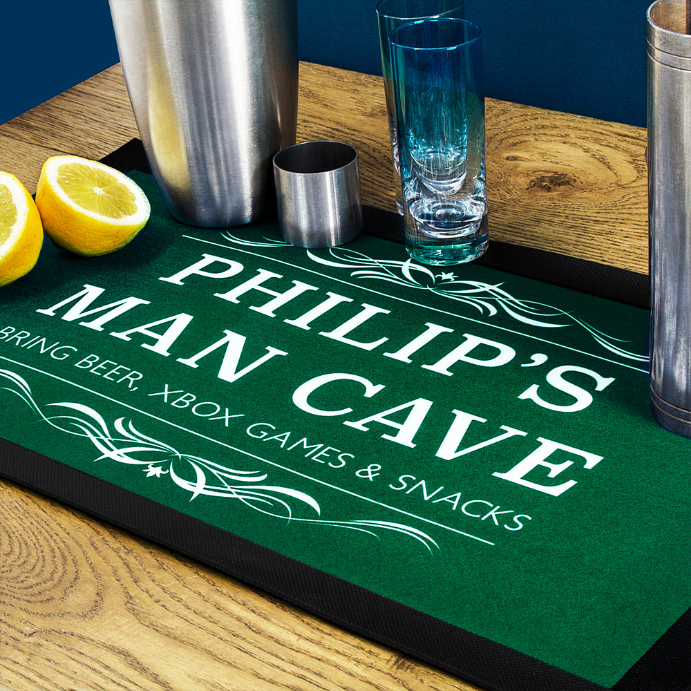 Personalized bar mat with 'PHILIP'S MAN CAVE' text on a wooden surface with bar accessories.