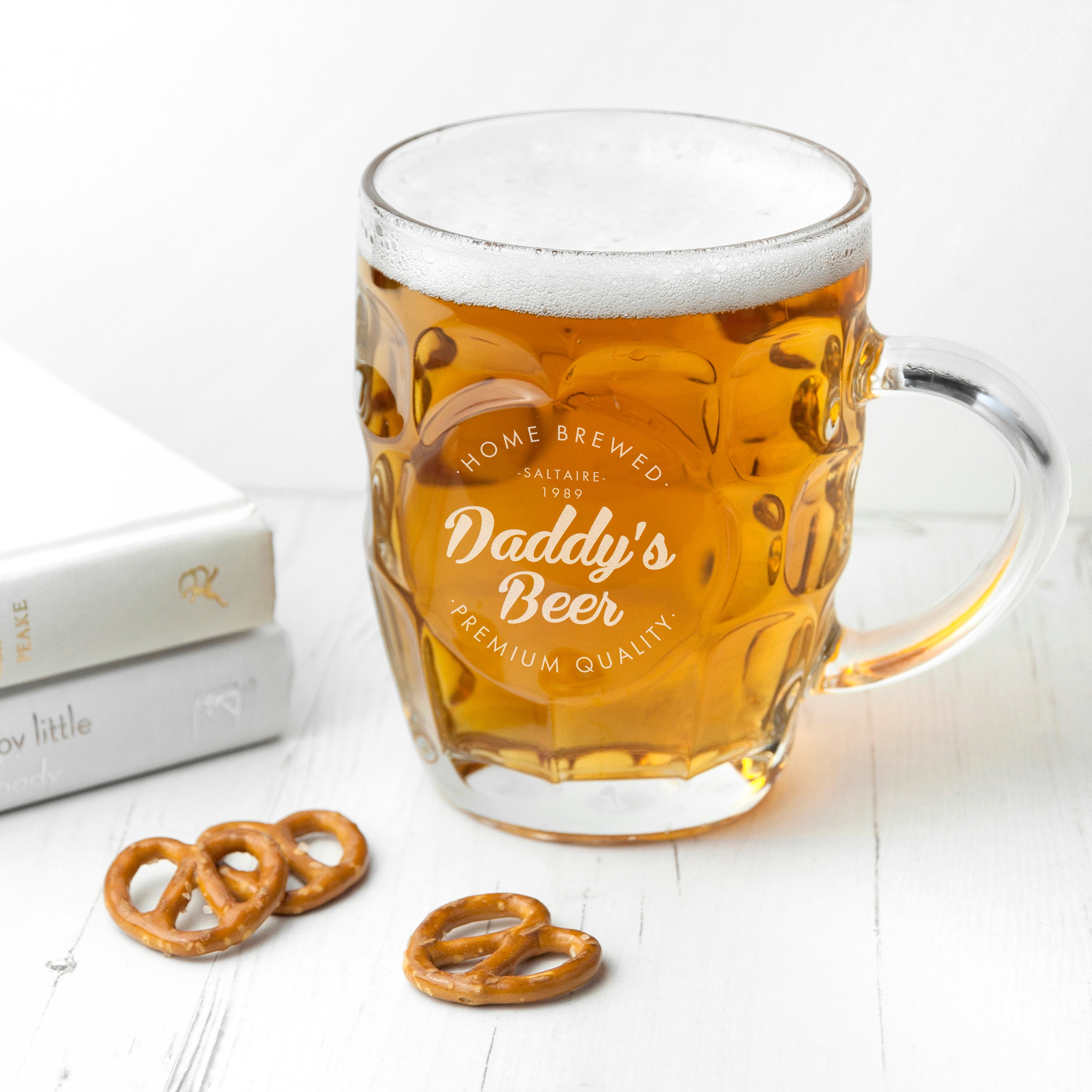 Glass mug with 'Daddy's Beer' engraving filled with beer on a white surface with pretzels.