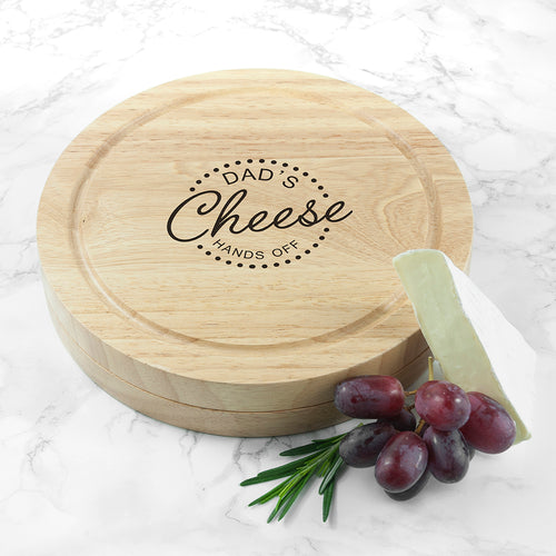 Wooden cheese board with 'Dad's Cheese Hands Off' engraving on a marble surface with grapes and a wedge of cheese.