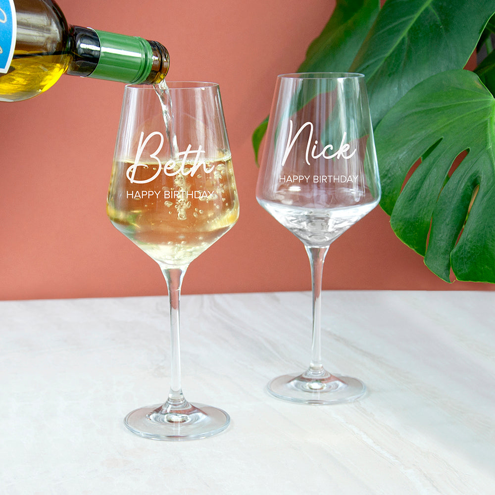 Two personalized wine glasses being filled with white wine against a pink background.
