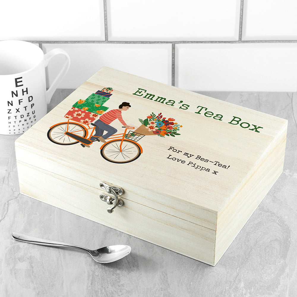 Wooden tea box with bicycle and flowers illustration, personalized text, on a tiled surface with a spoon.