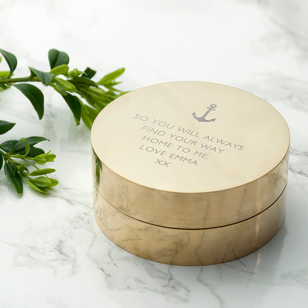 Round brass box with engraved text on a marble surface with green leaves.