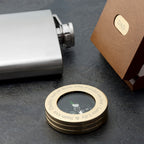 Brass compass with engraved text on a dark surface, next to a silver flask and brown box labeled 'DAD'.