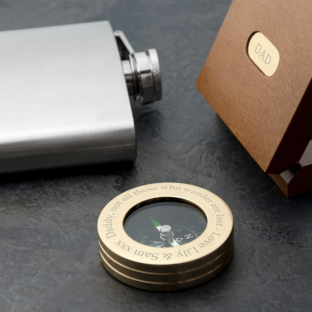 Brass compass with engraved text on a dark surface, next to a silver flask and brown box labeled 'DAD'.