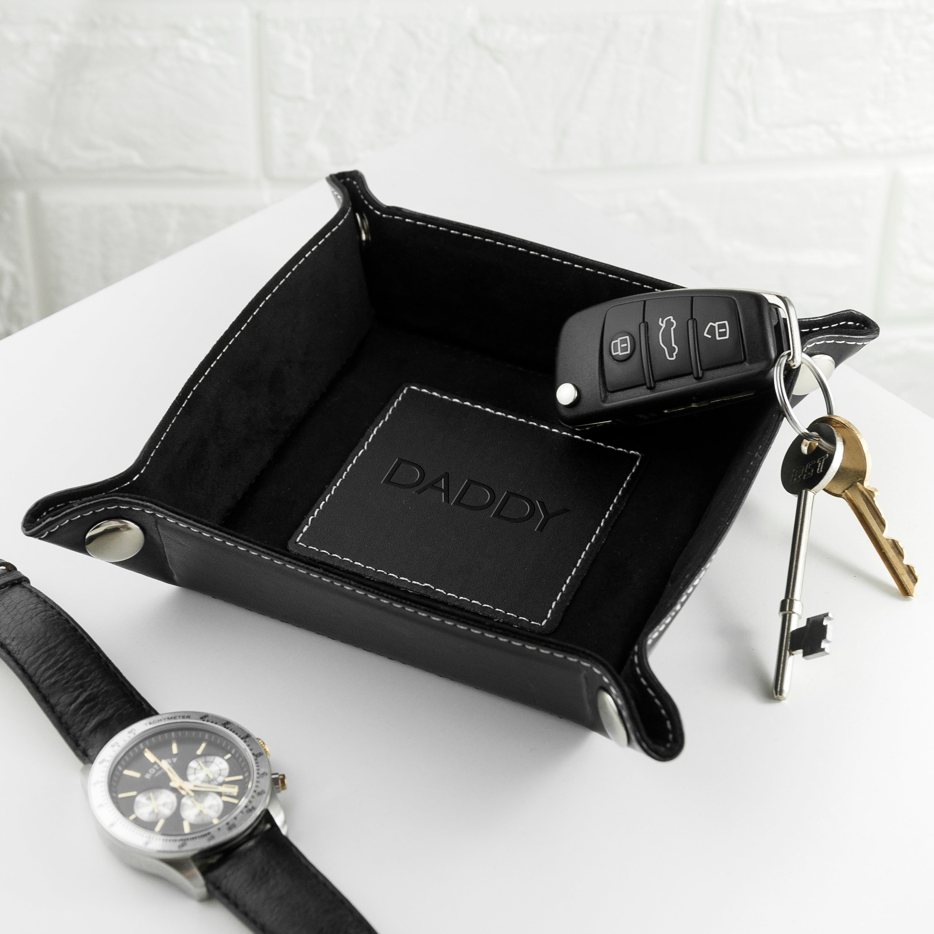 Black leather tray with 'DADDY' engraving, keys, and a watch on a white surface.