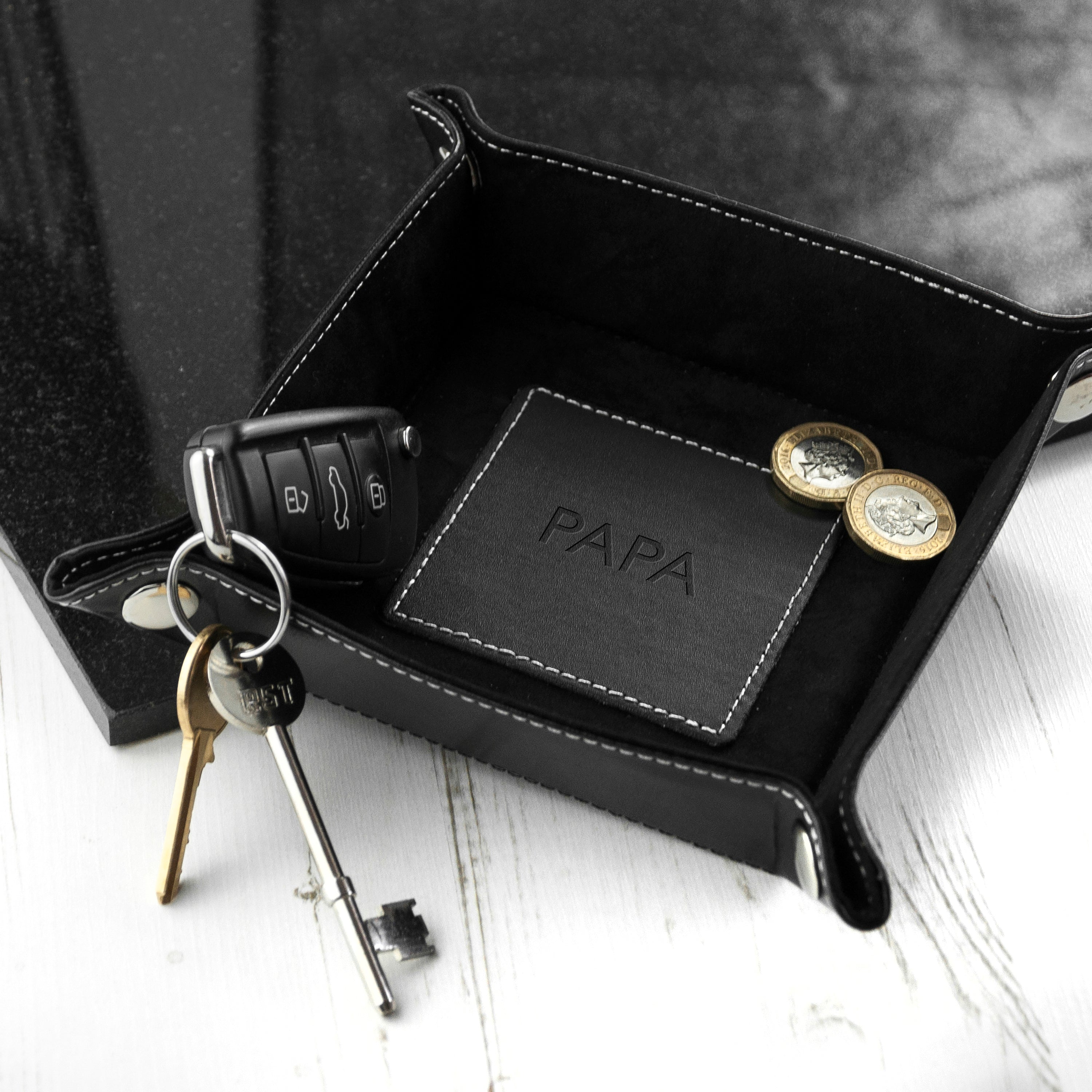Black leather tray with car keys, wallet, and coins on a white surface