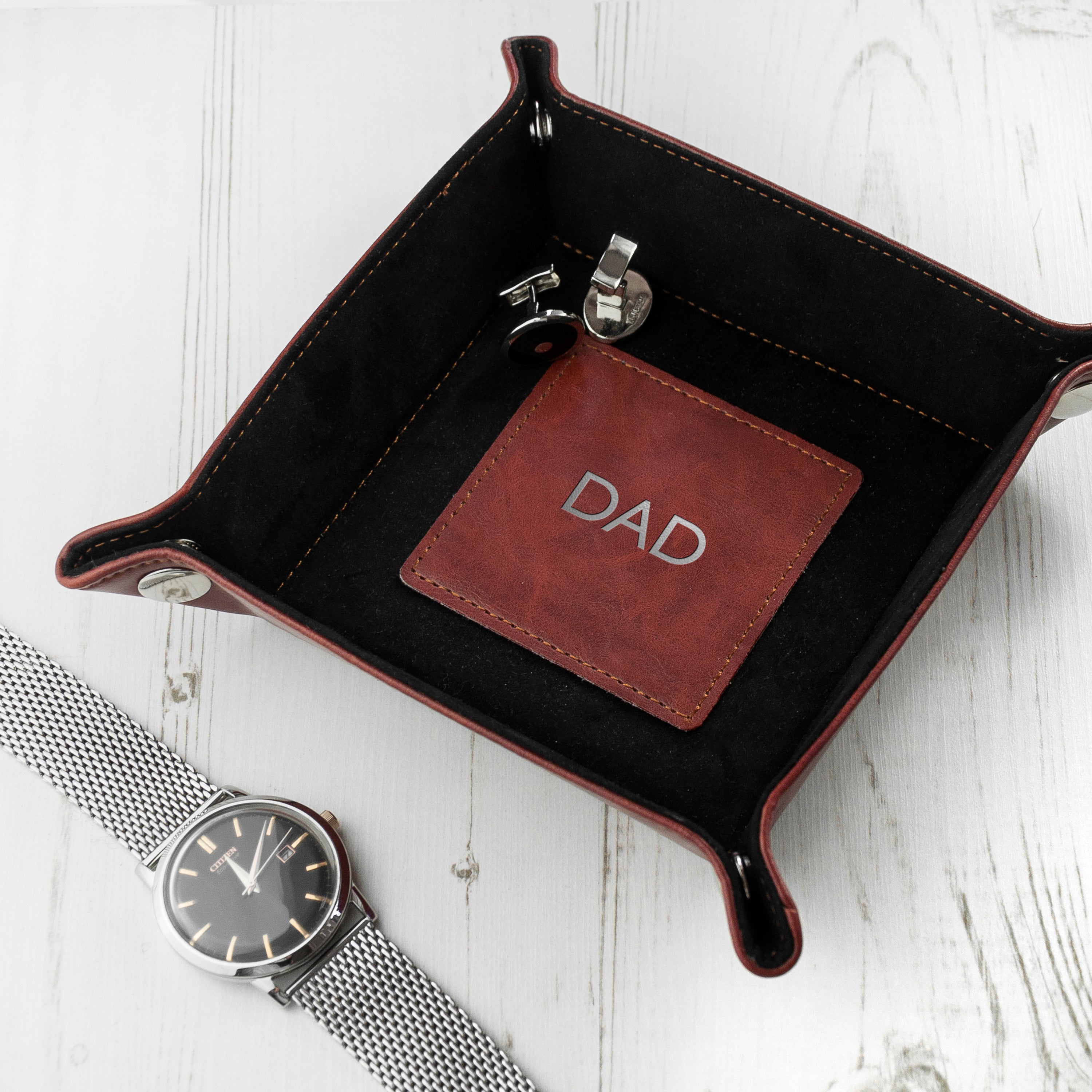 Black leather tray with a brown card labeled 'DAD' on a white wooden surface with a silver watch.