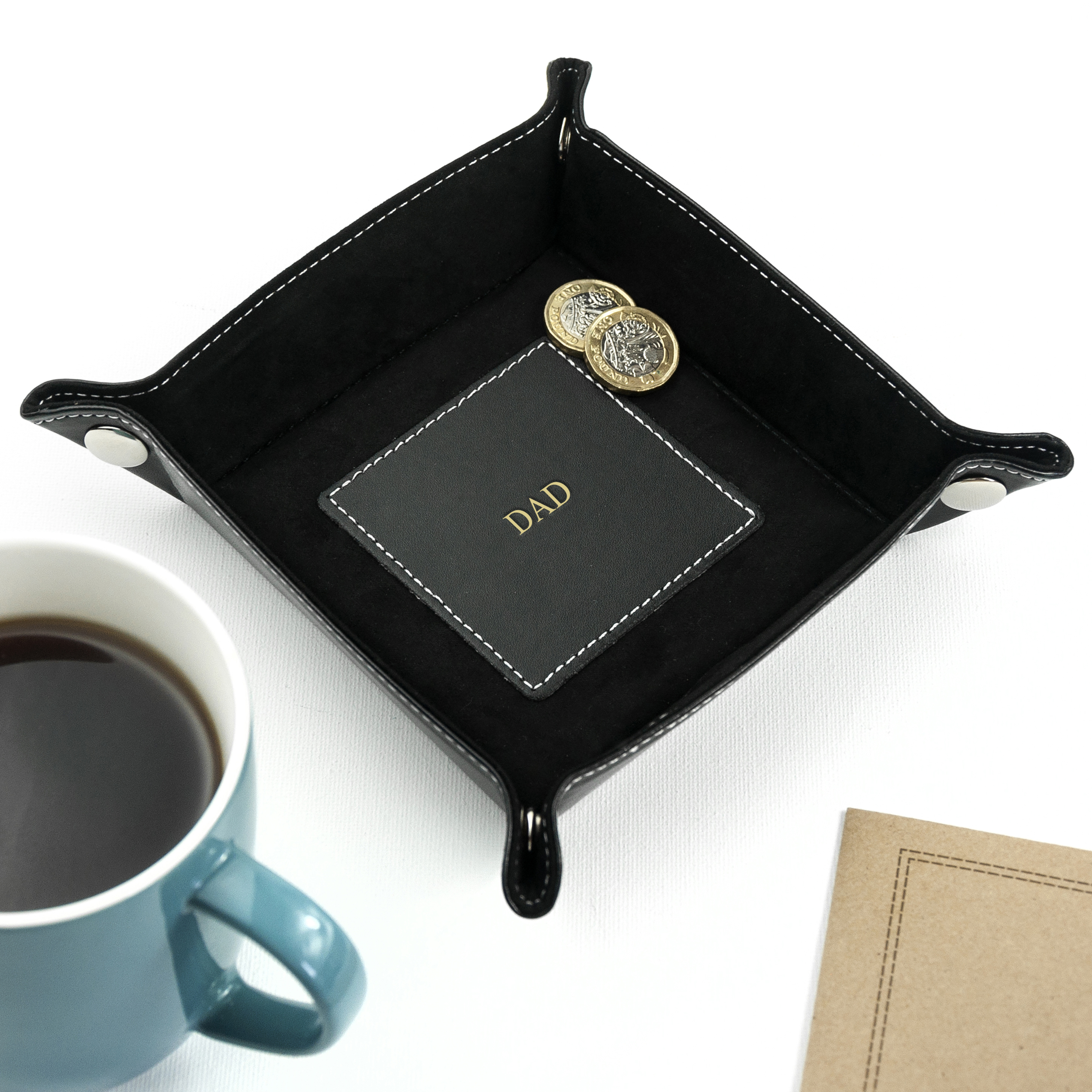 Black leather tray with 'POPS' branding, mug of coffee, and notebook on a white background