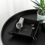Black leather tray with a watch, cufflinks, and a small plant on a white surface.