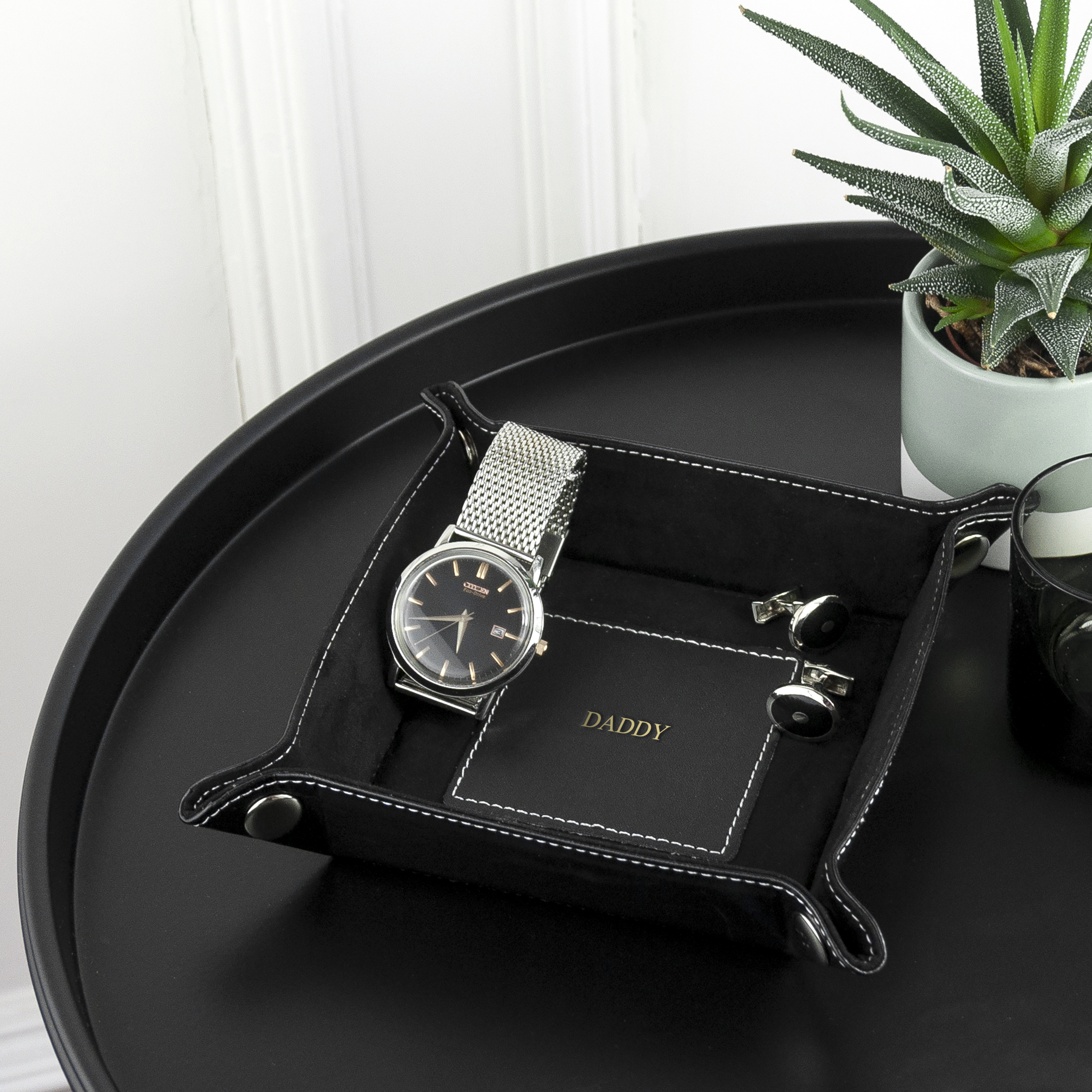 Black leather tray with a watch, cufflinks, and a small plant on a white surface.