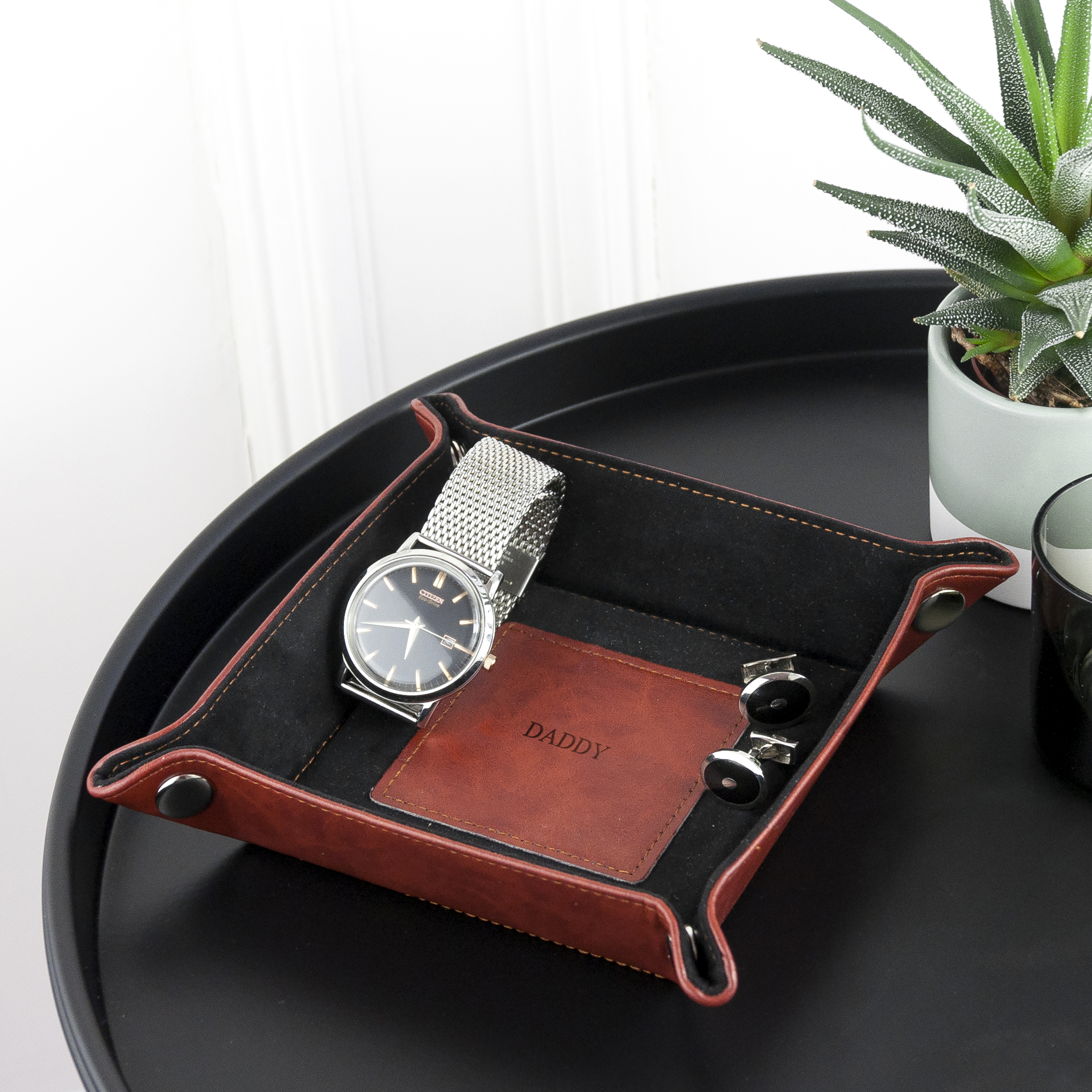 Brown leather tray with watch and card holder on a black tray with a plant in the background
