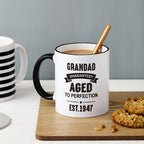 White mug with black text 'Grandad Guaranteed Aged to Perfection Est. 1947' on a wooden coaster with cookies and a striped mug.