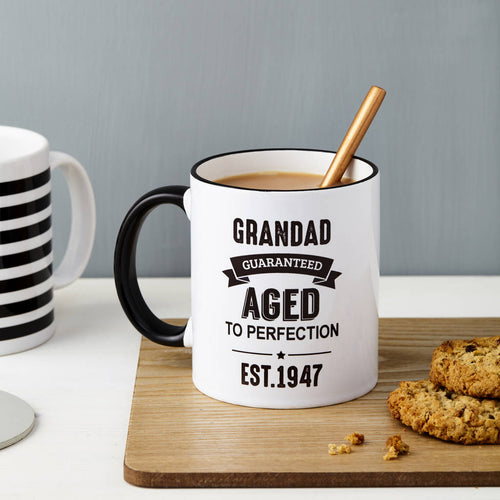 White mug with black text 'Grandad Guaranteed Aged to Perfection Est. 1947' on a wooden coaster with cookies and a striped mug.
