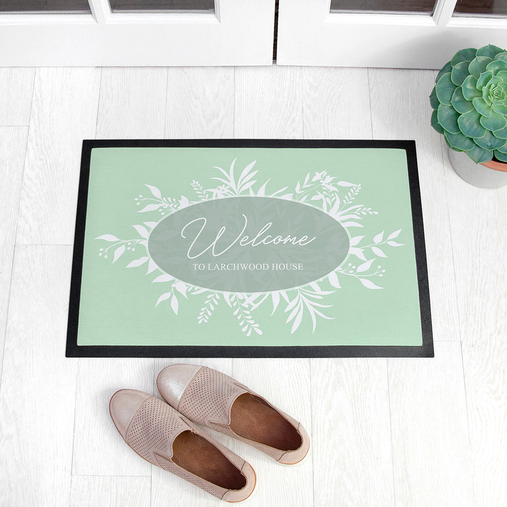 Doormat with 'Welcome to Larchwood House' text on a light wooden floor with shoes and a plant.