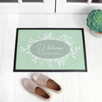 Doormat with 'Welcome to Larchwood House' text on a light wooden floor with shoes and a plant.