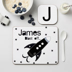 Placemat with a rocket design and 'James Blast off' text on a table with a bowl of cereal and blueberries.
